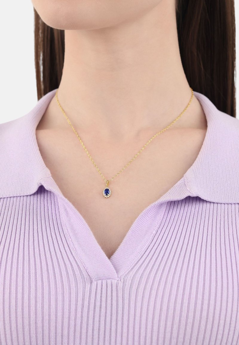 Collana in oro con un pendente di zaffiro circondato da piccoli diamanti, indossata da una persona con un top a righe di lavanda con colletto.
