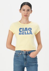 Yellow cropped t-shirt with "CIAO BELLA" printed in bold blue letters. Short sleeves and a round neckline. Paired with high-waisted blue jeans.