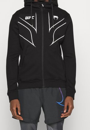 Mand iført sort UFC hoodie med hvide kantede striber og logoer, kombineret med mørke sportsshorts med en blå grafik på venstre ben.