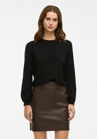 Must have pikkade varrukatega must pluus, ümar kaelus ja elastsed mansetid, kombineeritud sobiva pruuni kunstnahast mini-seeliku jaotises. Sile tekstuur, minimaalne disain.