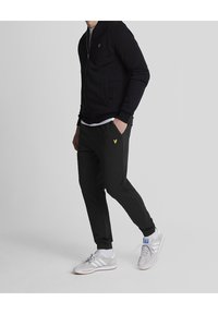Sort zip-up sweatshirt med ribbede manchetter, parret med sorte joggers med et gult logo. Båret med hvide og grå træningssko.