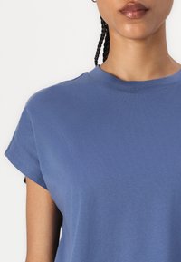 Blaues T-Shirt aus glattem Baumwollmaterial, mit Rundhalsausschnitt, kurzen aufgerollten Ärmeln und einer lockeren Passform, ohne sichtbare Muster oder Designs.