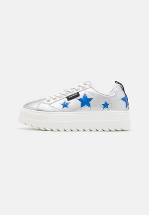 Zapatilla blanca con plataforma, puntera plateada, acentos de estrellas azules en los lados, etiqueta negra con la palabra "Hugo" y cordones blancos sobre un fondo liso.