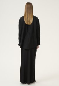 Camicia nera oversize con tessuto testurizzato, vestibilità ampia e maniche lunghe, abbinata a una gonna lunga nera, vista da dietro.