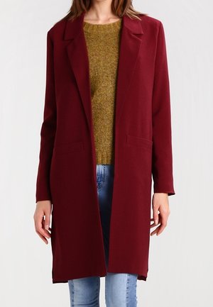 Femme portant un long manteau bordeaux par-dessus un pull moutarde et un jean bleu clair, debout devant un fond clair uni.