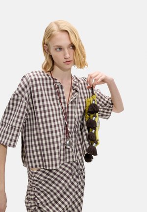 Kobieta ubrana w brązowo-biały top i spódnicę w kratę, trzyma żółtą torebkę z ciemnymi frędzlami i nosi długi naszyjnik z wisiorkiem.