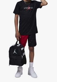 Camiseta negra con el logo "Jordan" en blanco y rojo, pantalones cortos rojos y negros, mochila negra con detalles en blanco, y zapatillas blancas con detalles texturizados.