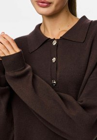 Jersey de punto marrón con cuello, que presenta cuatro botones decorativos y puños acanalados. Textura suave y corte relajado.