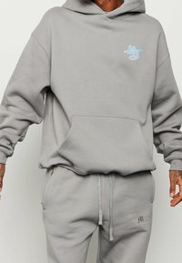 Sudadera gris con capucha y bolsillo tipo canguro, con un pequeño logo azul. Está hecha de un material suave, tiene un diseño relajado y puños acanalados.