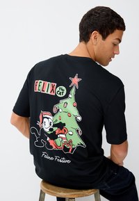 Črna majica z motivom Felixa mačka, ki okrašuje zeleno božično drevo z okraski, darili in besedilom "Mačja praznična".