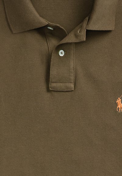 Nærbilde av brun poloskjortekrage og knappestolpe med en liten oransje brodert logo av en hest og rytter på høyre side av brystet.