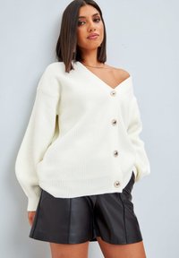 Krämfärgad ribbad cardigan med långa ärmar, v-ringning och fem stora knappar fram. Kombineras med svarta lädershorts.