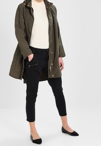 Olivgrön parkasjacka med huva, med dragkedja fram och fickor, bärs över en krämfärgad tröja och svarta avsmalnande byxor med dragkedjor.