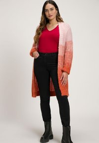 Neulecardigan, jossa on vaaleanpunaisen ja oranssin väriä vaihteleva sävy, löysällä istuvuudella ja ribbitetty pinnalla. Yhdistetty punaisen V-pääntien toppiin ja mustiin farkkuihin.