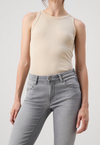 Top sin mangas beige hecho de una tela suave, combinado con jeans gris ajustados que cuentan con un cierre de botón y bolsillos frontales.