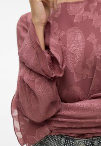 Blusa rosa polveroso con dettagli floreali testurizzati, maniche semi-trasparenti e vita cinchata. Indossata con una cintura nera decorata.