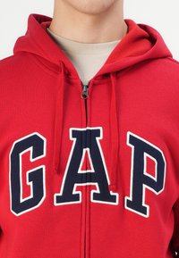 Rode zip-up hoodie met een marineblauwe "GAP" logo. Gemaakt van zachte stof met een fleece-gevoerde capuchon en drawstrings. Standaard pasvorm.