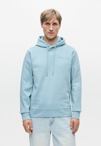 Ανοιχτό μπλε φούτερ φτιαγμένο από μαλακό ύφασμα, με μπροστινή τσέπη, κορδόνια και ένα διακριτικό λογότυπο στο στήθος. Συνδυάζεται με ανοιχτού χρώματος τζιν παντελόνι.