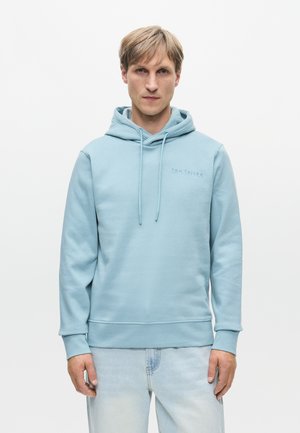 Ανοιχτό μπλε φούτερ φτιαγμένο από μαλακό ύφασμα, με μπροστινή τσέπη, κορδόνια και ένα διακριτικό λογότυπο στο στήθος. Συνδυάζεται με ανοιχτού χρώματος τζιν παντελόνι.