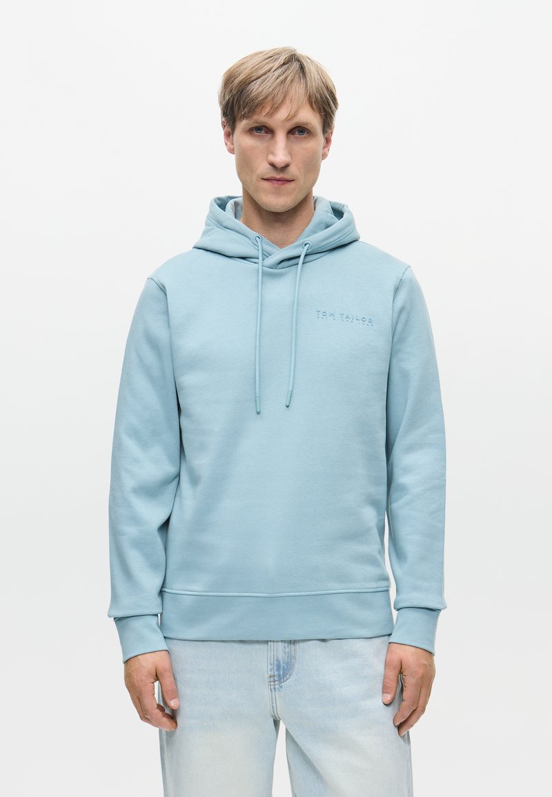 Ανοιχτό μπλε φούτερ φτιαγμένο από μαλακό ύφασμα, με μπροστινή τσέπη, κορδόνια και ένα διακριτικό λογότυπο στο στήθος. Συνδυάζεται με ανοιχτού χρώματος τζιν παντελόνι.