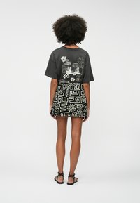 Woman with curly hair stands facing away, wearing a black graphic t-shirt with summer-themed prints and a black patterned mini skirt.