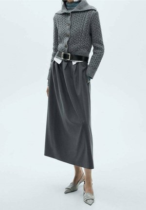 Personne portant un cardigan gris en maille torsadée, une longue jupe plissée taille haute avec une ceinture noire et des escarpins slingback à bout pointu.