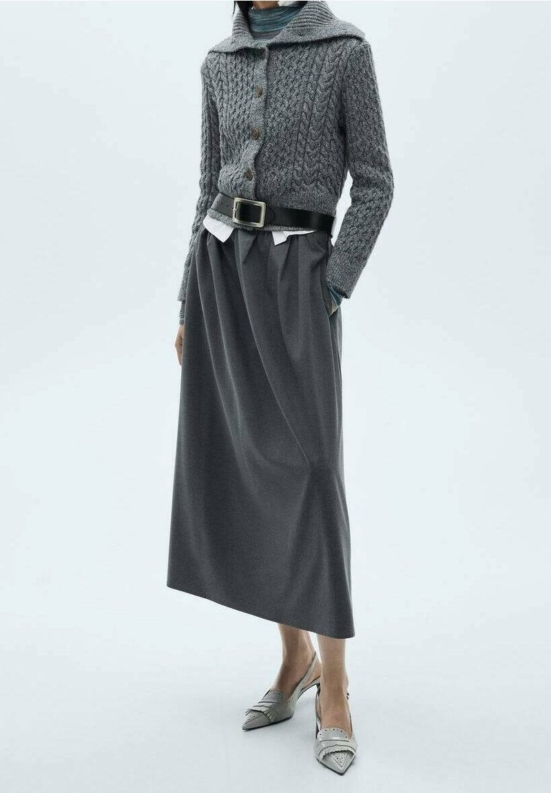 Personne portant un cardigan gris en maille torsadée, une longue jupe plissée taille haute avec une ceinture noire et des escarpins slingback à bout pointu.