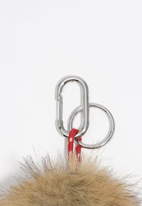 Llavero de piel sintética con un pompón color tostado, clips de metal plateado y una correa trenzada roja. Presenta una mezcla de texturas y formas circulares.