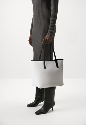 Personne portant une robe grise côtelée et des bottines noires à talons, tenant à côté d'elle un grand sac fourre-tout blanc avec des poignées noires.