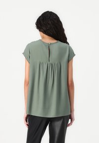 Frau mit langen schwarzen Haaren, die eine lockere, kurzärmlige Bluse in Salbeigrün mit Knopfleiste hinten und schwarze Hose trägt, mit dem Rücken zur Kamera.