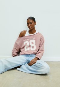 Felpa oversize rosa con la scritta "LOS ANGELES 28" in bianco, abbinata a jeans baggy azzurri. Tessuto morbido, vestibilità rilassata.