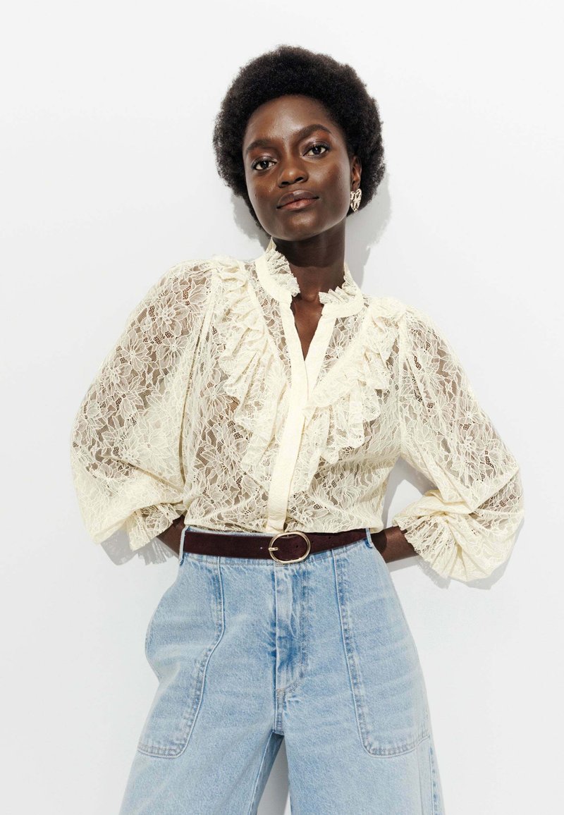 Blouse en dentelle crème avec des volants et un col haut, associée à un jean en denim taille haute bleu clair et une ceinture marron foncé.