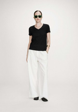 Femme portant un t-shirt noir à col en V, un pantalon blanc à jambes larges, des chaussures noires, des lunettes de soleil vertes et des bracelets en or, debout avec une main dans la poche.