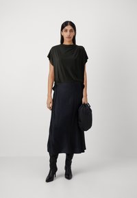 Černý top s krátkým rukávem v kombinaci s dlouhou tmavou sukní; hladká, splývavá látka; modelka drží malou kulatou černou kabelku.