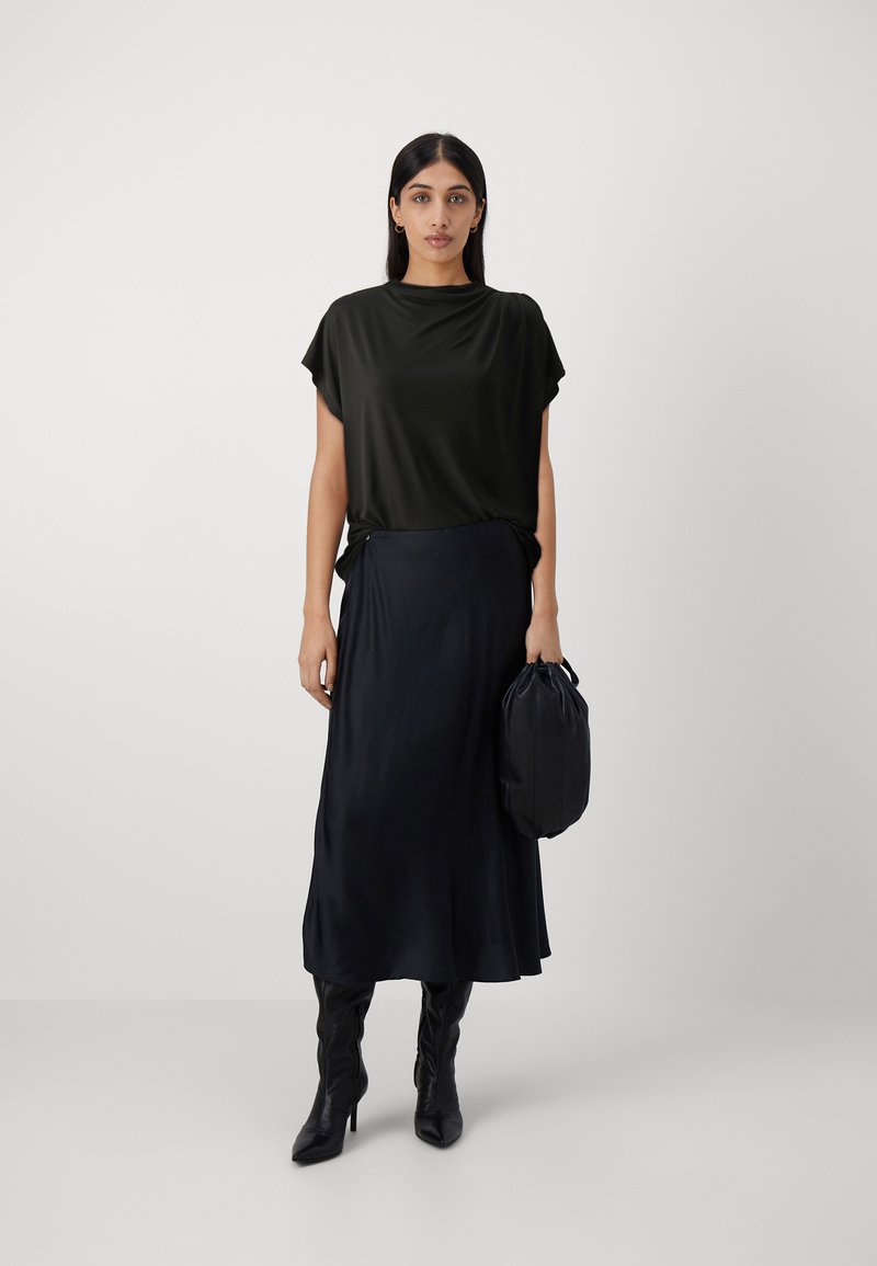 Schwarzes, kurzärmeliges Oberteil kombiniert mit einem langen, dunklen Rock; glattes, fließendes Material; Model hält eine kleine, runde schwarze Tasche.