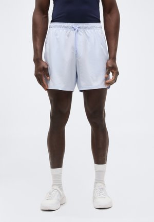 Férfi modell világoskék zsinóros rövidnadrágot, fehér zoknit, fehér tornacipőt és sötét ujjatlan felsőt visel, karjai lazán lefelé engedve áll.