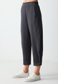 Dunkelgraue Hose mit elastischem Bund, verkürztem Design, zwei Seitentaschen und einem Knopfakzent am Saum. Kombiniert mit weißen Sneakern.