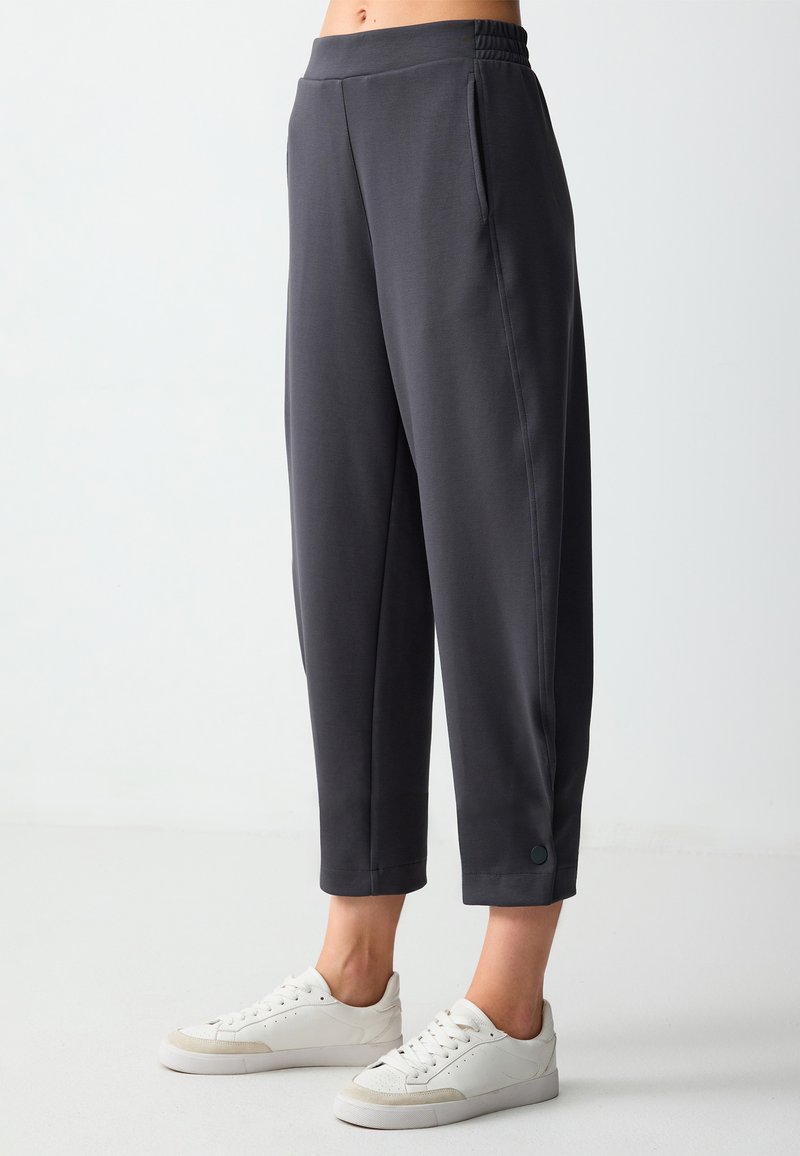 Dunkelgraue Hose mit elastischem Bund, verkürztem Design, zwei Seitentaschen und einem Knopfakzent am Saum. Kombiniert mit weißen Sneakern.