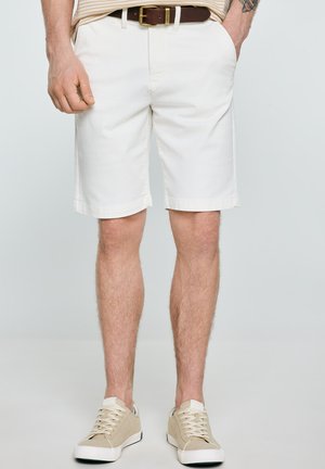 Mężczyzna ubrany w białe krótkie spodenki do kolan, beżowe trampki i brązowy pasek, stoi z jedną ręką w kieszeni, a drugą zrelaksowaną.