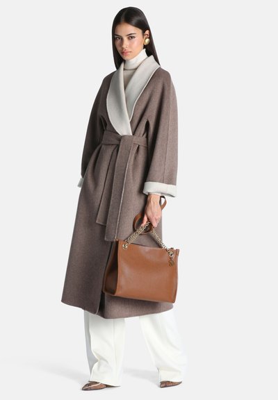 Femme portant un long manteau marron ceinturé avec des poignets blancs, un pantalon blanc et tenant un sac à main en cuir marron avec des poignées en chaîne dorée.