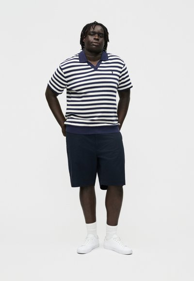 Bărbat stând cu mâinile la spate, purtând un polo cu mâneci scurte cu dungi albastre închis și albe, pantaloni scurți albaștri închiși, șosete albe și încălțăminte sport albă.