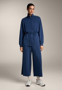 Mornarsko modra prevelika jakna z zadrgo in visokim ovratnikom, z vrvico v pasu, v kombinaciji z širokimi hlačami. Gladka tkanina z sproščenim krojem in minimalnimi detajli.