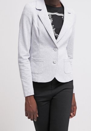 Femme portant un blazer gris clair ajusté avec deux boutons et des poches plaquées sur une chemise noire à motifs et un pantalon noir.