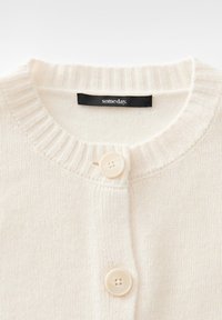 Creme farbener Strickcardigan mit geripptem Rundhalsausschnitt, zwei cremefarbenen Knöpfen und einem schwarzen Etikett mit der Aufschrift "someday." innen am Kragen.