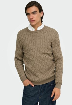 Jeune homme portant un pull beige à maille torsadée par-dessus une chemise blanche à col et un jean foncé, debout avec une main dans la poche.
