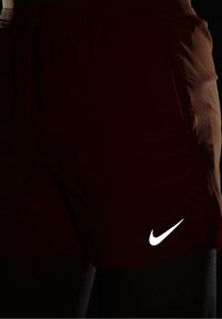 Nike urheilushortsit tummasta punaisesta kankaasta, jossa on sileä pinta. Oikeassa alakulmassa valkoinen logo ja elastinen vyötärönauha, jossa on kiristysnauha.