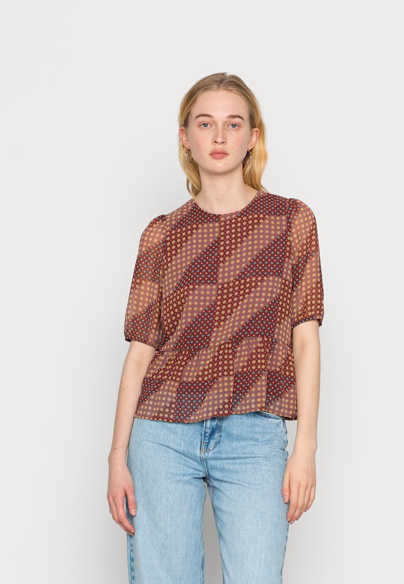 Blusa transparente em ferrugem e creme com padrões geométricos, mangas curtas bufantes, decote redondo e uma bainha solta e franzida. Combinada com jeans claros.