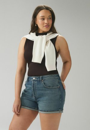 Nő fekete ujjatlan felsőt és kék farmerrövidnadrágot visel, fehér pulóvert terítve a vállára egy egyszerű szürke háttér előtt.