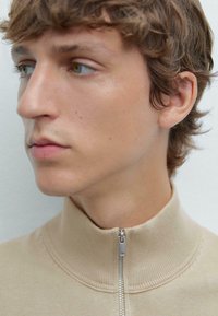 Beiger Reißverschluss-Pullover mit hohem Kragen, gestrickter Textur und metallischem Reißverschluss. Nahaufnahme eines Modells, das natürliche Hauttöne und welliges Haar zeigt.
