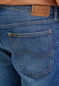 Ciemnoniebieskie dżinsy z denimu z brązową skórzaną metką z napisem "Lee". Posiadają żółte przeszycia, tylną kieszeń oraz klasyczny krój.